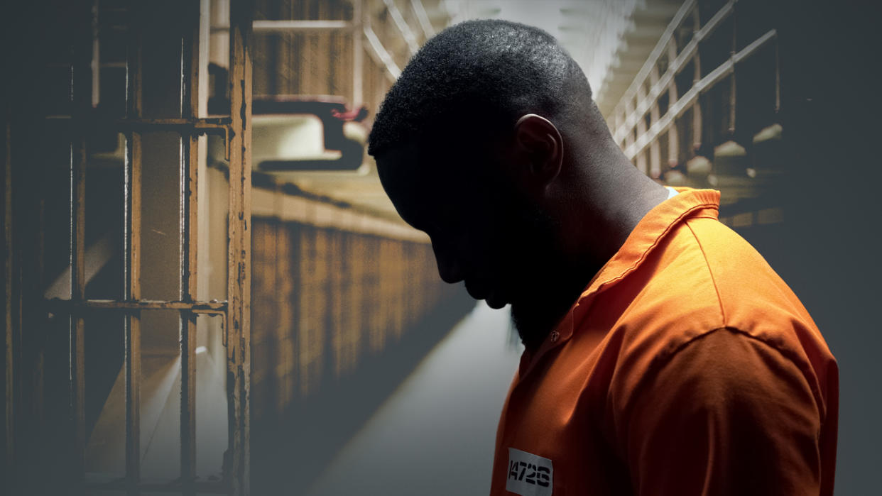 A Black man with his head down wearing an orange uniform is seen against a background showing the interior of a prison.