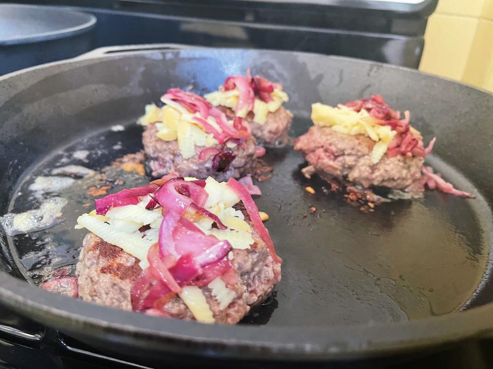 hamburgers cooking in a cast iron skillet with caramelized onions and cheese