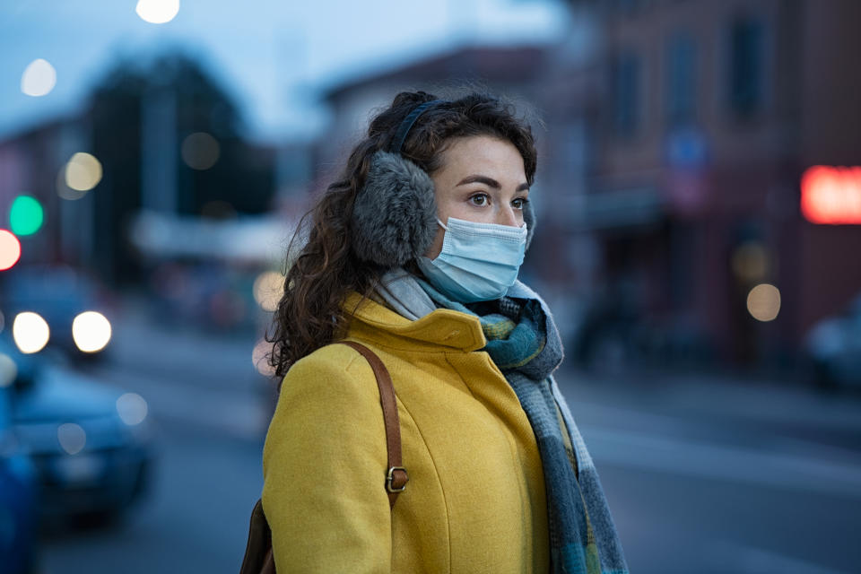 Woman wearing medical protective mask outdoor during dusk. Young woman wearing face mask against pollution standing outdoors on winter evening. Girl with gray earflap in winter city street during coronavirus outbreak.