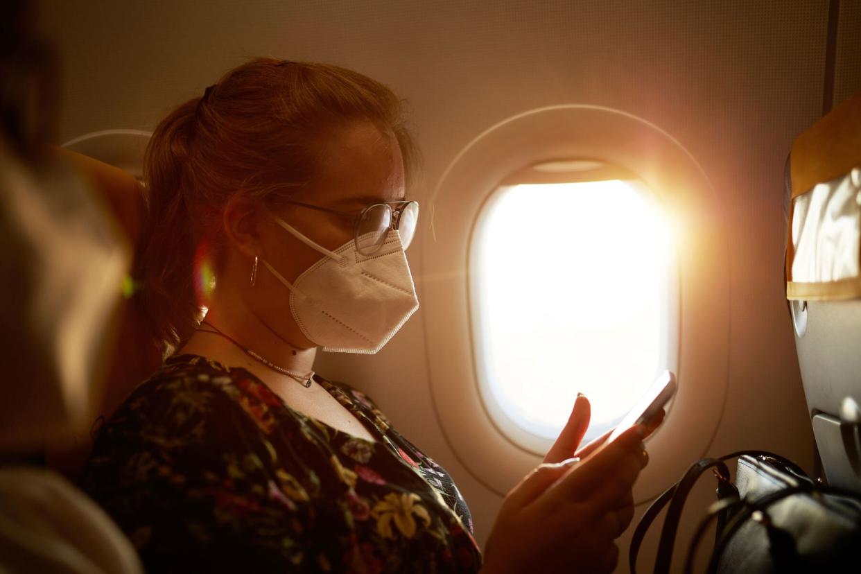 Side view of young adult wearing glasses and protective mask checking smart phone while sitting in airplane window seat in time of COVID-19.