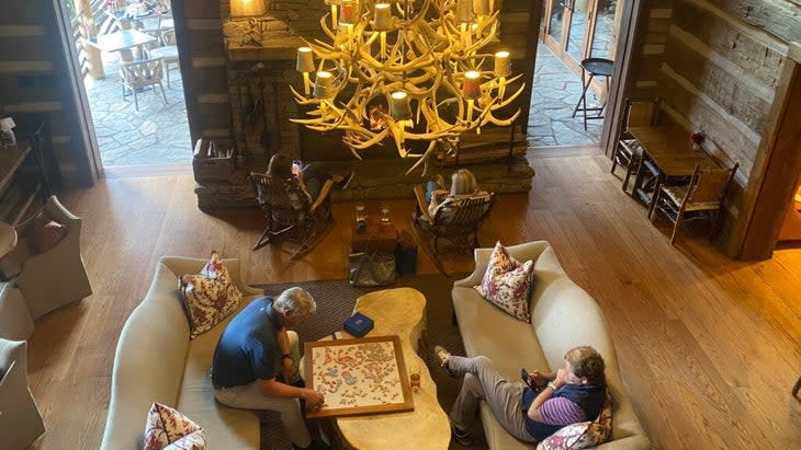 A couple, each sitting on their own sofa, ponder a puzzle on a table between them. A huge antler chandelier hangs overhead.