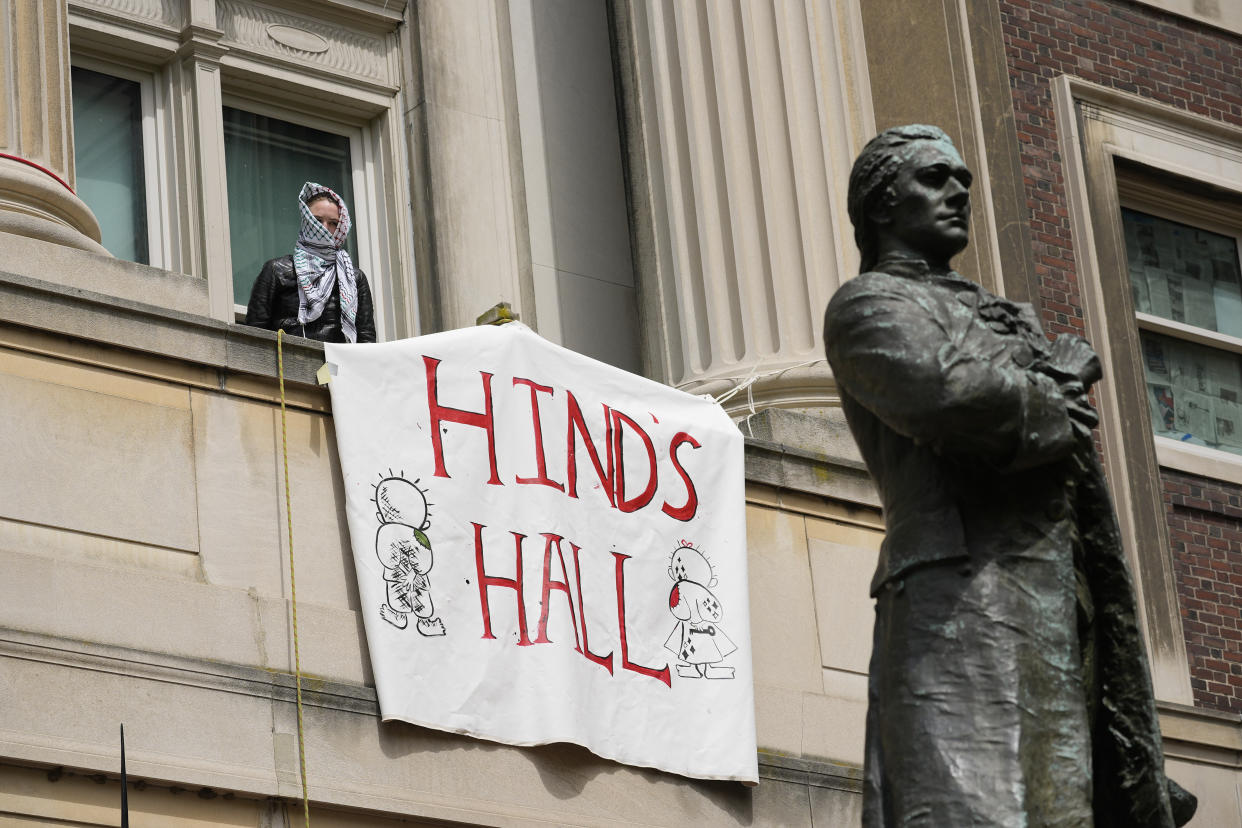 A person wearing a headscarf stands at a window above a sign reading: Hind's Hall