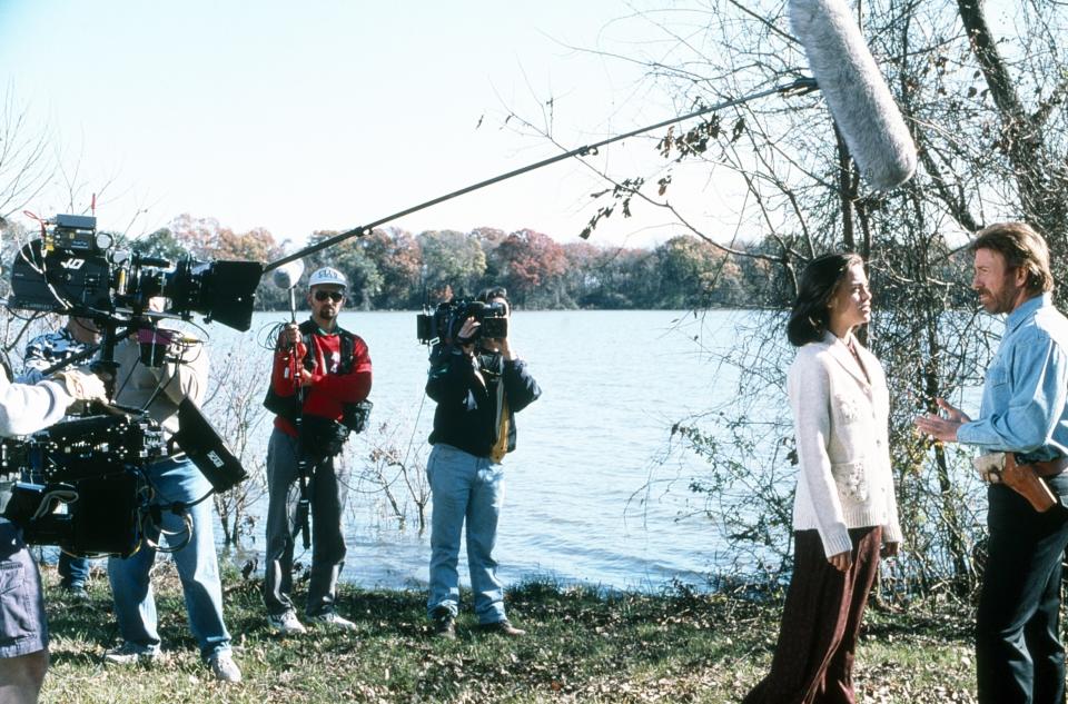 Chuck Norris films a scene by a lake, crew and camera equipment visible