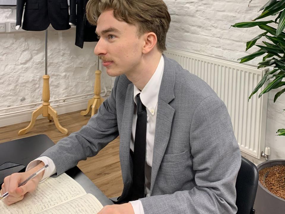 A reporter in a grey suit sits in a classroom and looks up, while writing in a notebook