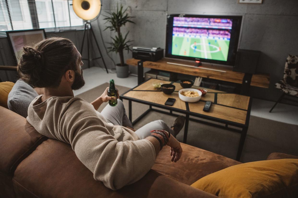 Young man sitting on sofa at home and watching soccer game. He is cheering for favorite soccer team.