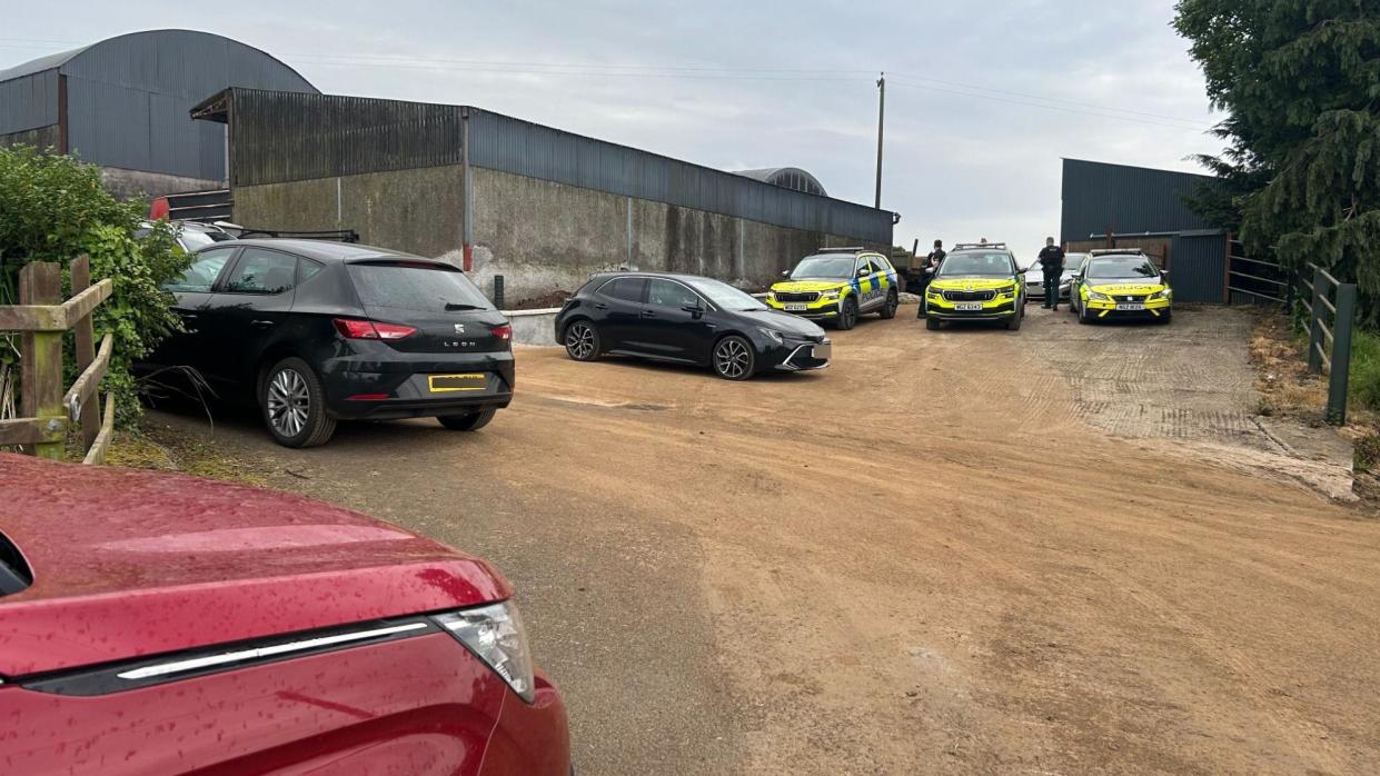 Cars and police vehicles sit on a dirt path near farm buildings