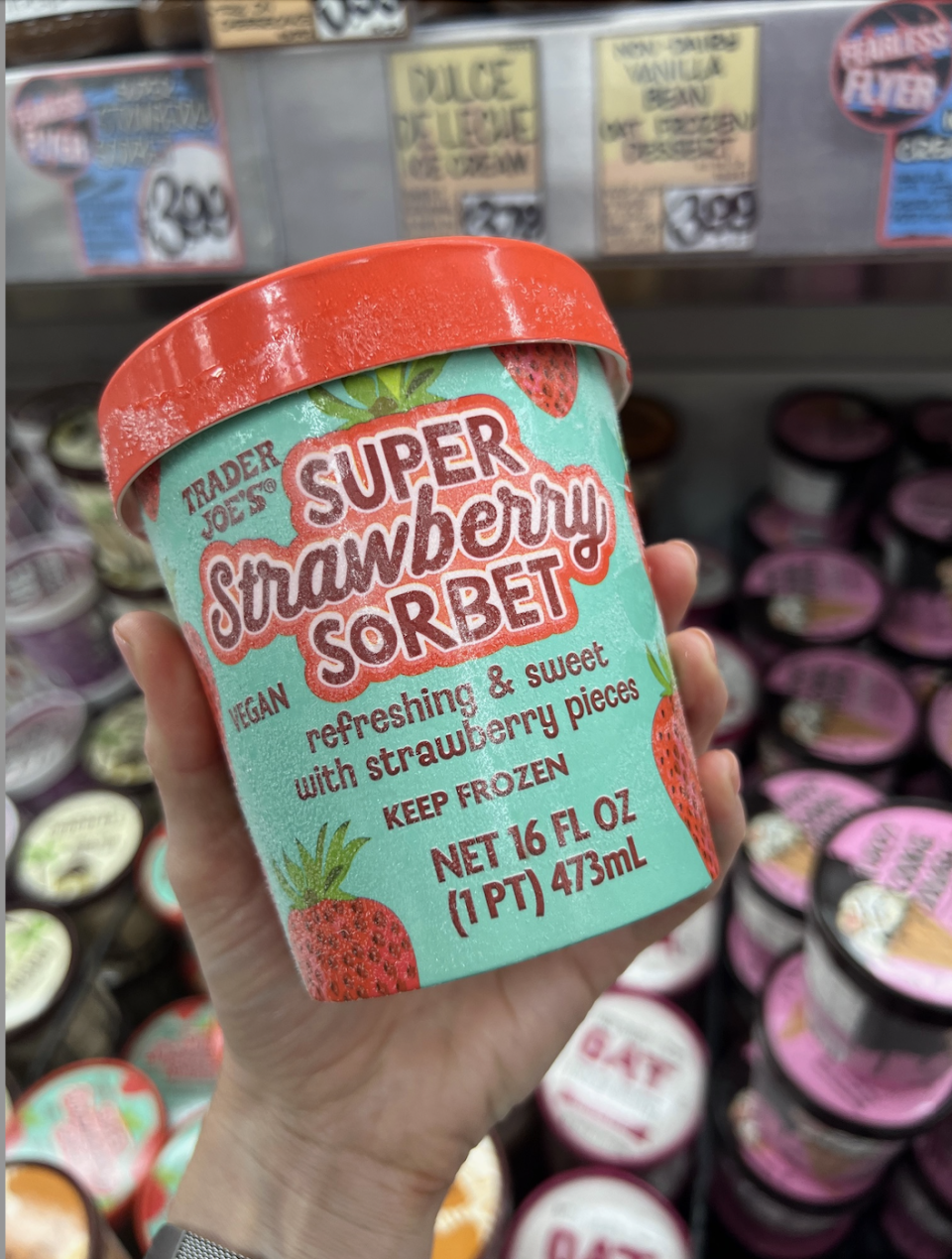 A hand holding a pint of Trader Joe's Super Strawberry Sorbet, vegan, 16 fl oz, in a store aisle with various other ice cream flavors in the background