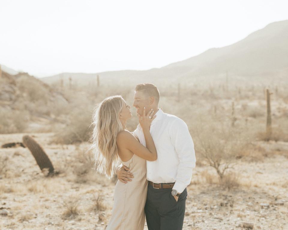 A woman touches a man's face as he hugs her in a desert.