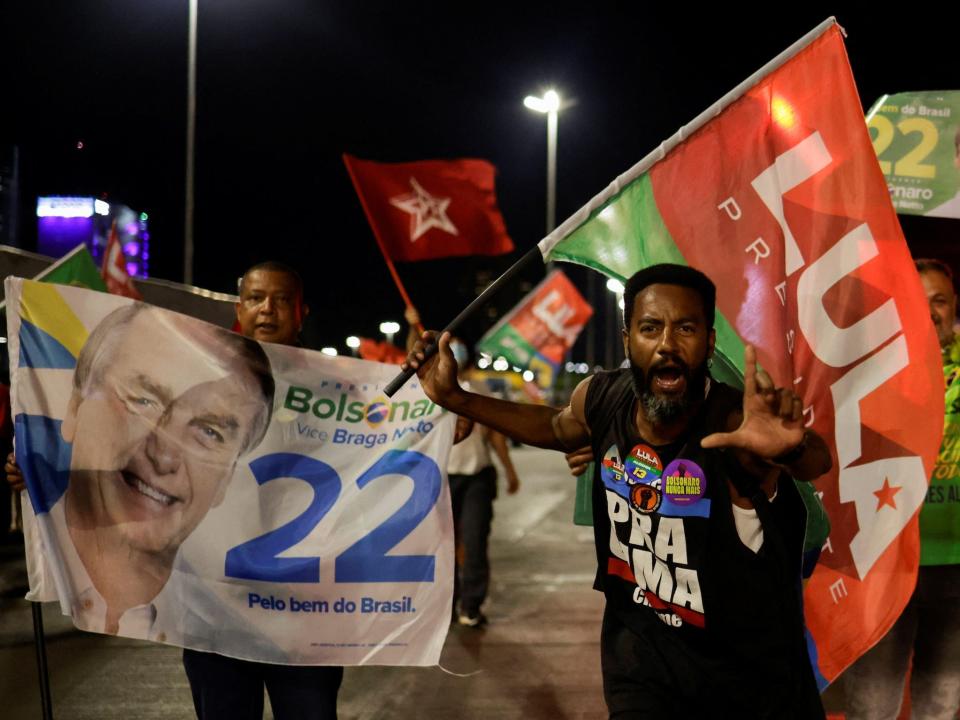 crowd of people waving flags some supporting jar bolsonaro some supporting Luiz Inacio Lula da Silva one man holding fingers in L shape at night