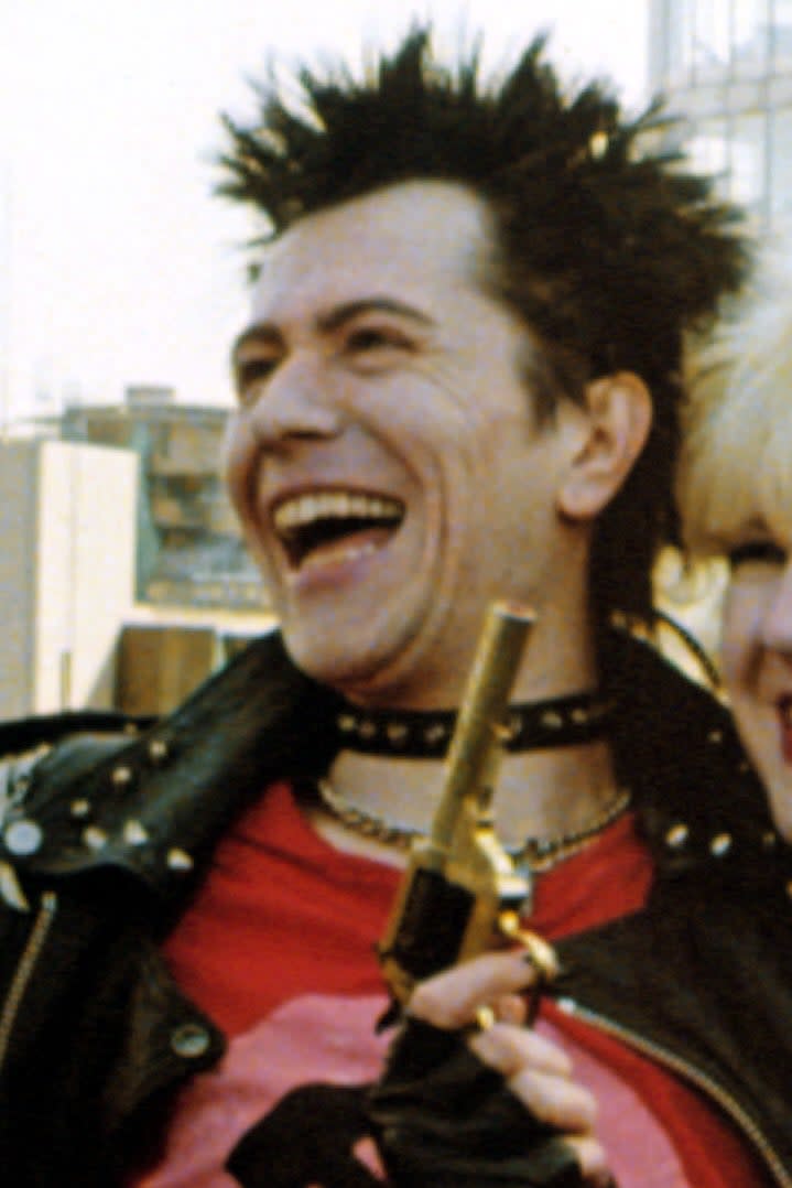 Two individuals, one with spiky hair and a leather jacket, and the other with curly blonde hair and a leather jacket. Both are smiling and holding a gun prop