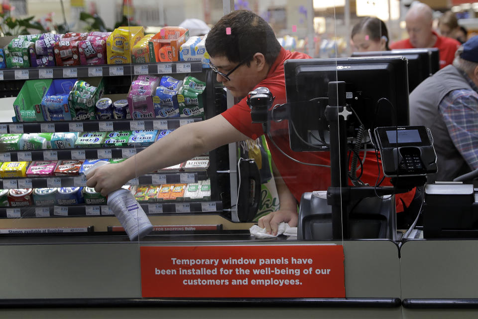 FILE - In this March 26, 2020, file photo, Garrett Ward sprays disinfectant on a conveyor belt between checking out shoppers behind a plexiglass panel at a Hy-Vee grocery store in Overland Park, Kan. From South Africa to Italy to the U.S., grocery workers — many in low-wage jobs — are manning the front lines amid worldwide lockdowns, their work deemed essential to keep food and critical goods flowing. (AP Photo/Charlie Riedel, File)