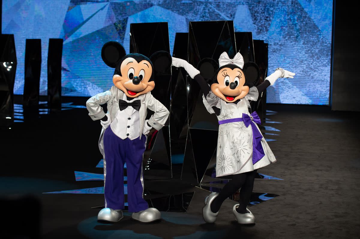 Two people in mouse costumes are standing in a room. The person on the left has their hands on their hips and is wearing a male costume: a silver shirt with a black button down, blue pants, and silver shoes. The person on the right is wearing a female costume: white dress with a blue sash tied aorund its waist, silver shoes, black tights, and a white ribbon. 