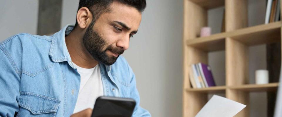 Young indian businessman holding phone reading bank receipt calculating taxes, ethnic man using smartphone mobile application checking bill document, managing money finances, loan expenses.