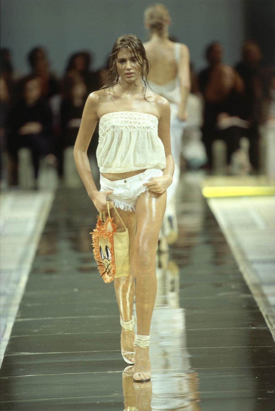 A model on the runway at the Spring/Summer 2000 Chloé ready-to-wear collection designed by Stella McCartney, wearing white tube top with smocking at top edge, fringed hot pants, high-heel sandals with white and gold ankle bands, and carrying a straw bag with cat-face design. (Photo: Getty Images)