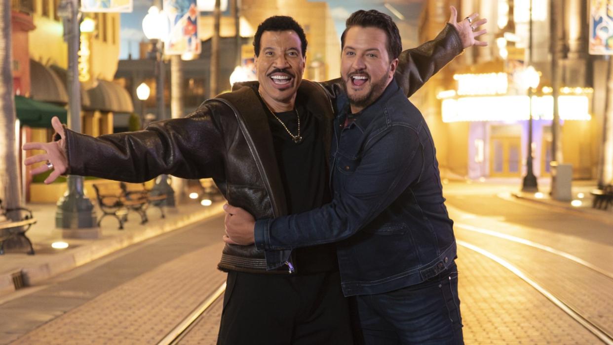  American Idol coach Lionel Richie stands with his arms outstretched in the middle of a street at night, with fellow judge Luke Bryan hugging him around the waist. 