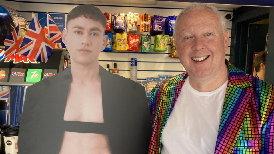 A smiling man dressed in a rainbow coloured blazer, standing next to a cardboard cut-out of Olly Alexander