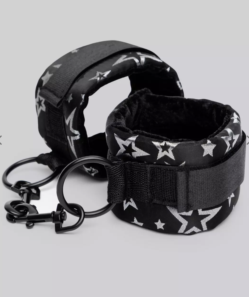 A black pair of Bondage Boutique Star Handcuffs with white star print and Velcro straps with short metal black chain.