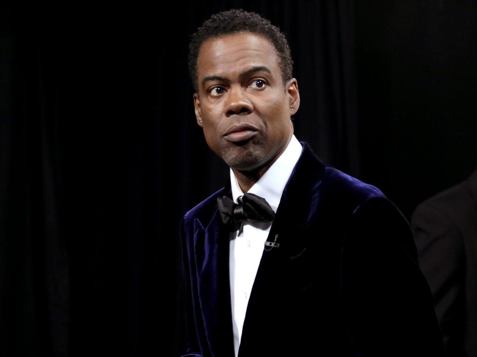 An image of Chris Rock wearing a tuxedo, carrying a red envelope
