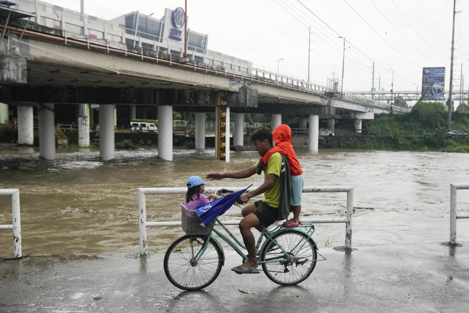 A man and children ride a bicycle beside a swollen river due to enhanced rains brought about by Typhoon Doksuri on Thursday, July 27, 2023, in Marikina city, Philippines. Typhoon Doksuri lashed northern Philippine provinces with ferocious wind and rain Wednesday, leaving several people dead and displacing thousands of others as it blew roofs off houses, flooded low-lying villages and triggered dozens of landslides, officials said. (AP Photo/Aaron Favila)