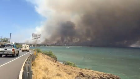 A plume of smoke spreads across the sky during a wildfire in Kihei, as vehicles move along a road in Maui