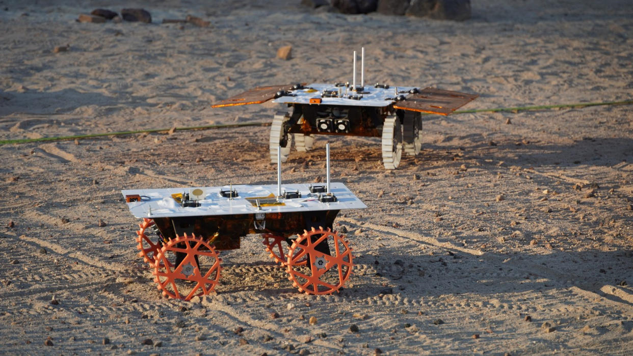  Two small four-wheeled robotic rovers drive in a sandy enclosure during a test. 