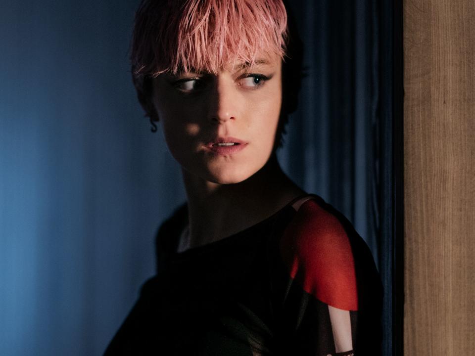 emma corrin as darby hart in a murder at the end of the world, with pink hair and looking around a corner