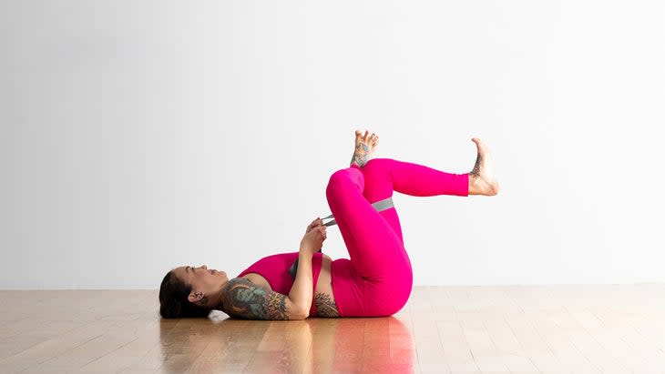 Woman practices a Pigeon Pose prep variation. She is on her back with both knees lifted toward her torso. Her right ankle is crossed over her left knee creating a figure-four. She has a strap around her right thigh and is holding it with both hands to pull it toward her.