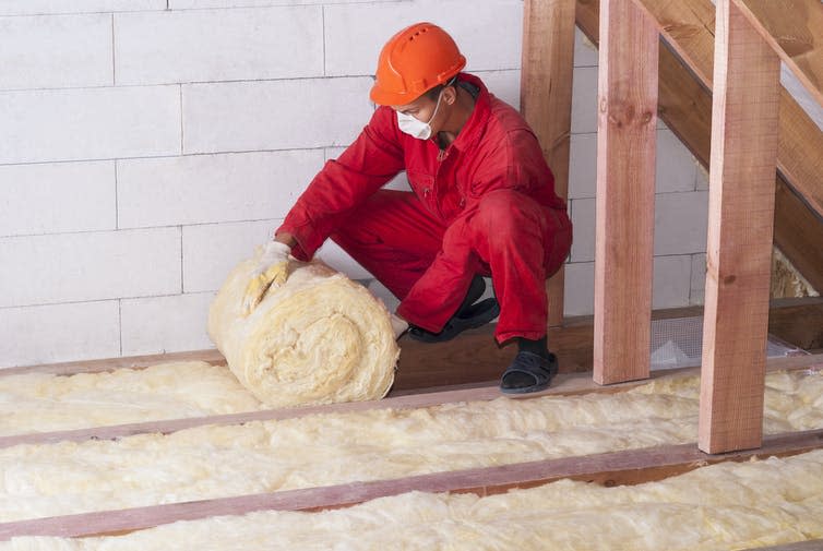 A worker in red overalls unrolls thermal insulation in a loft.