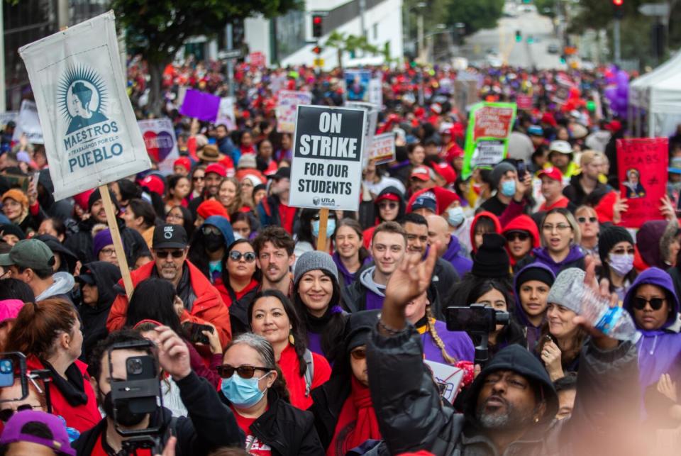 A crowd of protesters, many wearing red and purple. A picket sign says "On Strike For Our Students."