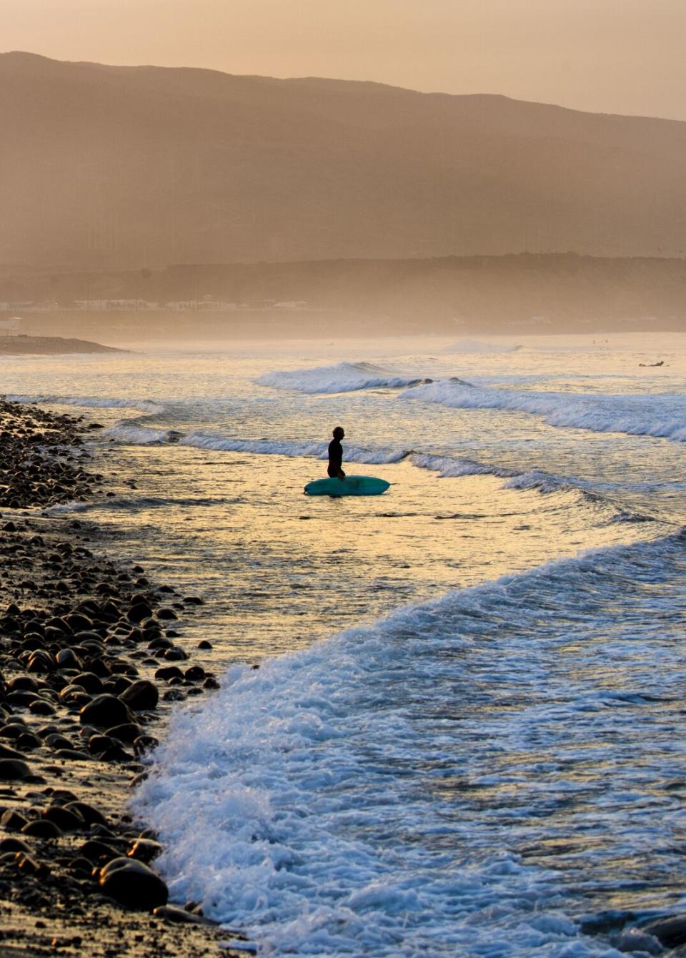 A lone surfer walks across the cobble-stones at sunrise to surf.