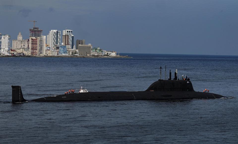A black Russian submarine surfaces above bluish ocean water with the cityscape of Havana, Cuba, in the background.