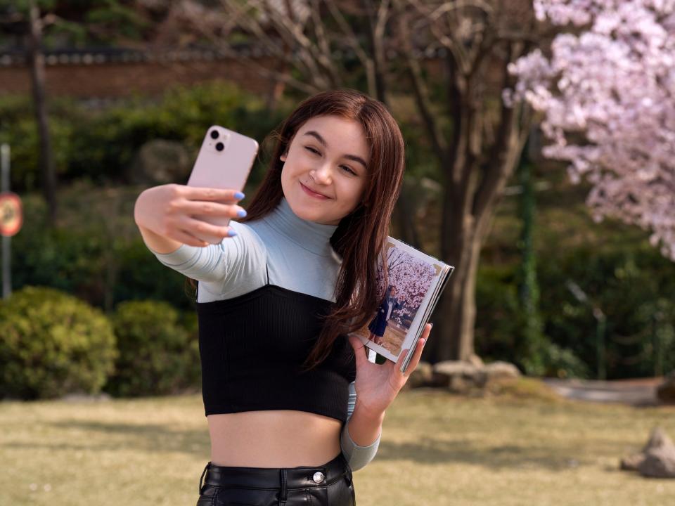 anna cathcart as kitty in xo kitty. she's a teenage girl wearing a blue turtleneck and cropped black tee, holding a photo of her mother in front of a cherry blossom tree. kitty snaps a selfie of herself, posing in front of the same tree.