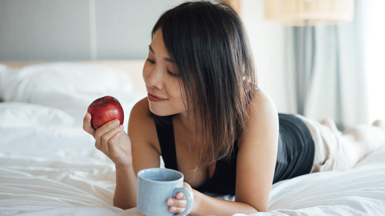  A woman with long dark hair lies in bed holding a red apple in one hand a cup of coffee in the other. 