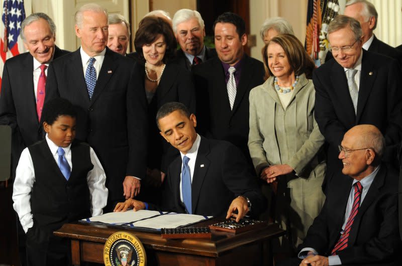 U.S. President Barack Obama signs the Affordable Care Act surrounded by Marcelas Owens (L) of Seattle, Vice President Joe Biden, Vickie Kennedy (behind the president), House Speaker Nancy Pelosi, Senate Democratic leader Harry Reid (R), Rep. John Dingell (R, seated) and other Congress members in the East Room of the White House in Washington, D.C., on March 23, 2010. File Photo by Pat Benic/UPI