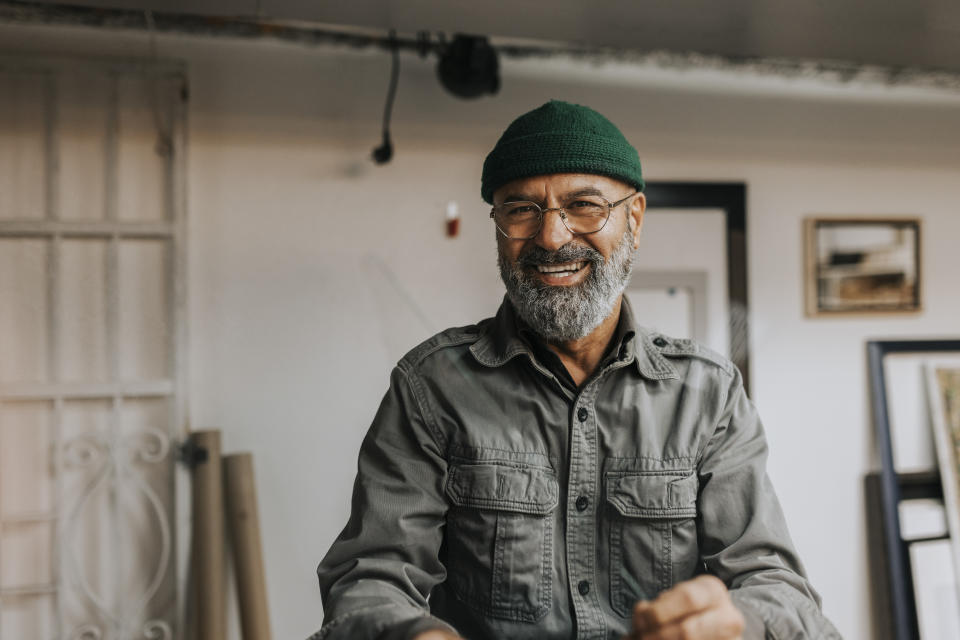 An older man with a beard wears a green beanie and glasses, smiling in a casual indoor setting