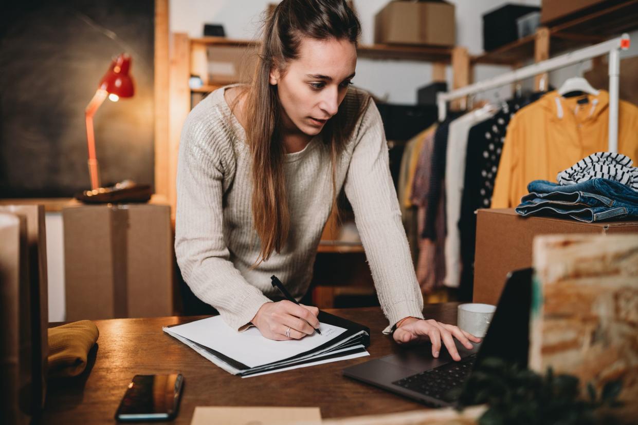 A young woman is checking her laptop and writing on her notebook. She is the owner of an online clothing chop.