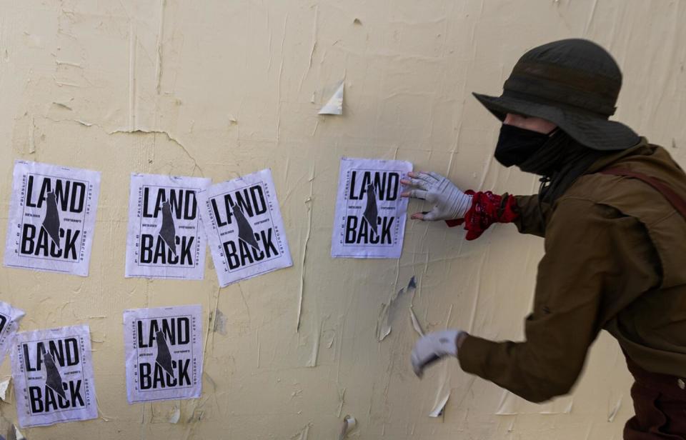 A person wearing a dark hat and mask has one hand on a white flier with the words Land Back on a white wall