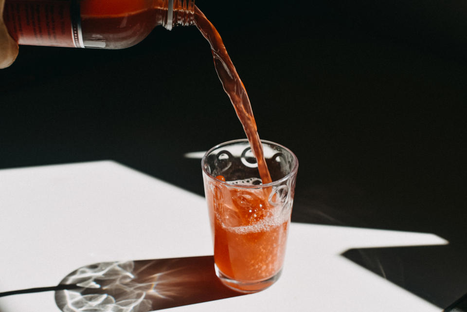 A clear glass being filled with a fizzy orange beverage poured from a bottle. The glass casts a shadow on a white surface