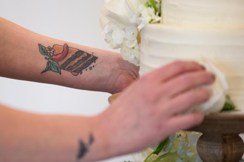 Whitney Leighton 's cake tattoo is seen as she decorates a cake in flowers at 1868 Farmhouse Wedding and Event Venue. COVID-induced parosmia has made her work more challenging than ever, but she continues baking for weddings, birthdays and other events.