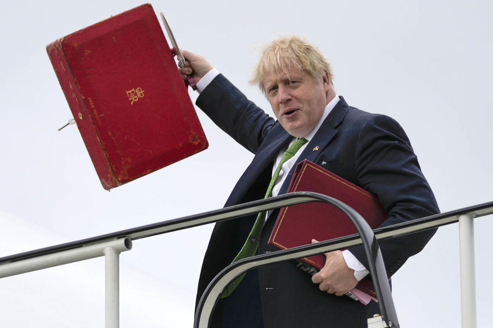Boris Johnson waves with a hand that also holds a red satchel while holding bound material under his other arm as he apparently descends an airplane gangplank.