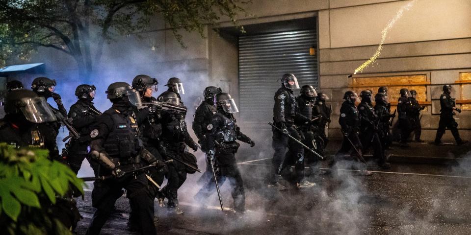 Portland police officers wearing riot gear and holding batons march forward during a protest.