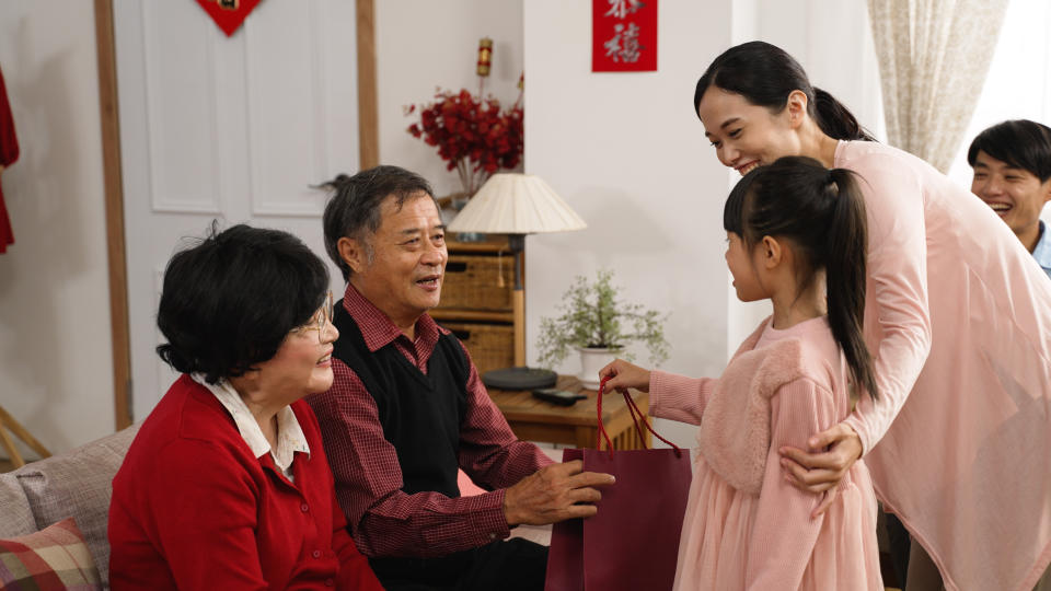 asian little girl and her mother in light red visiting grandparents with gift bag during spring festival. happy elderly man praising and patting on granddaughter’s shoulders