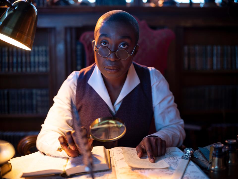 vivienne acheampong as lucienne in the sandman. she's wearing a smart black vest over a white collared shirt and is looking skeptically over a pair of round glasses while sitting at a busy desk