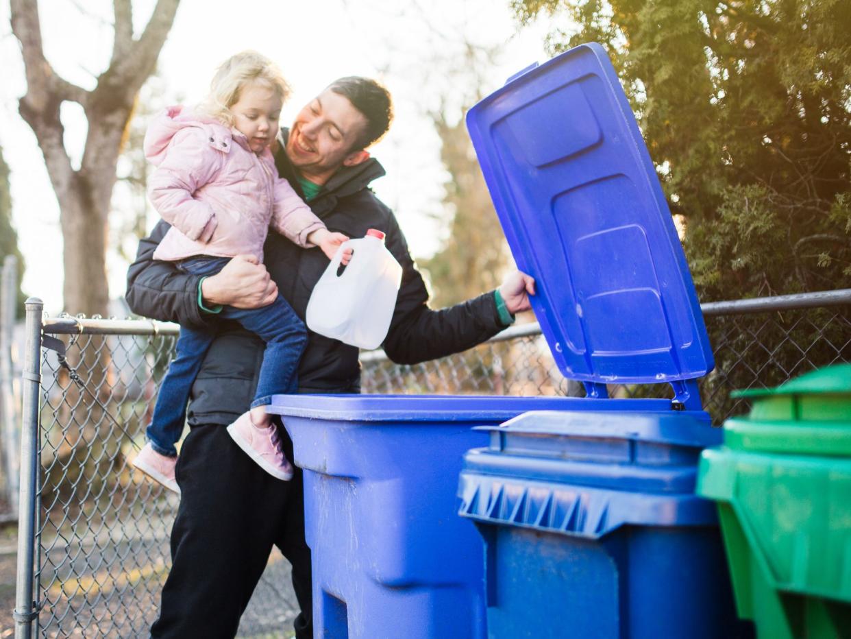 A dad holds his daughter as he teaches her about the environmentally friendly practice of recycling plastic and cardboard waste. A good practical learning opportunity.
