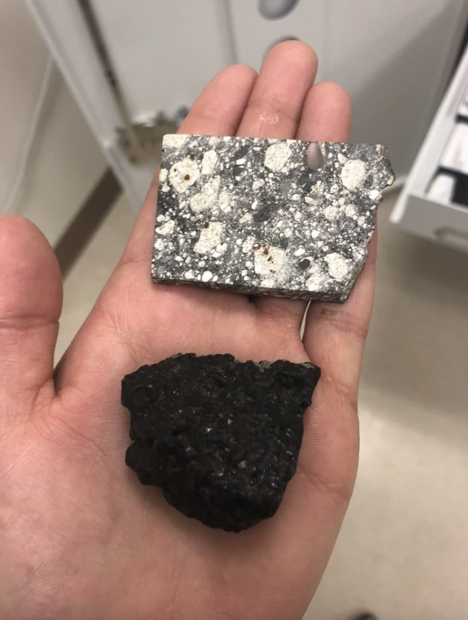 A person's hand holding a speckled square mineral sample above a rough, irregular-shaped black sample