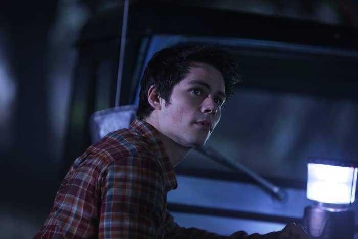 Dylan O'Brien as Stiles Stilinski in a plaid shirt, looking concerned, with a flashlight in a dimly lit scene from Teen Wolf