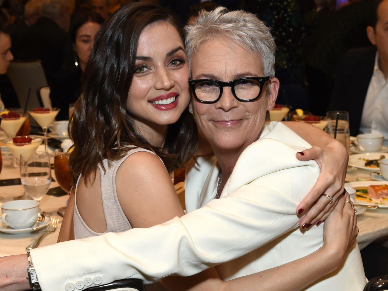 Ana de Armas, smiling in a tan sleeveless dress, sits with her arms around Jamie Lee Curtis, seated beside her in a white suit and black-rimmed glasses, for a photo op during a dinner ceremony.