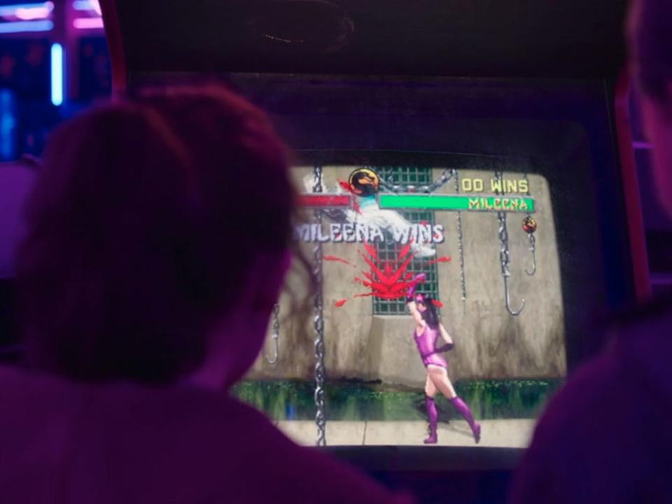 ellie and riley in the last of us, seen from behind, playing mortal kombat in an arcade. they're illuminated by purple and blue light