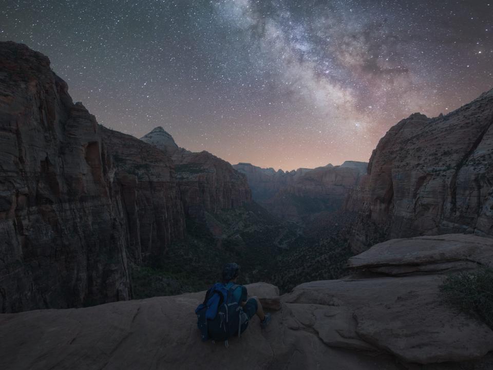 A person wearing a backpack sits on rocks looking at many stars in the Milky Way at Zion National Park