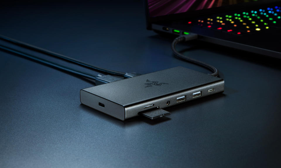 Product marketing photo of the Razer USB C Dock. The hub sits on a desk with a gaming laptop behind it. It has several open ports and an SD Card halfway out. It sits on a dark blue desk with dramatic shadows.